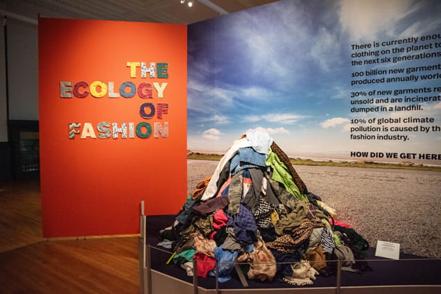 A photo of a pile of clothes next to the wall reading "The Ecology of Fashion."