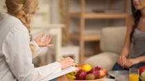 Woman in lab coat speaking with another woman, a bowl of fruit sits on a table between them