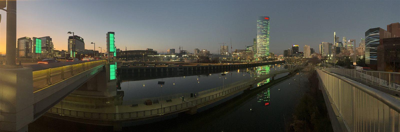 A panoramic view of the Philadelphia skyline at sunset showing buildings and objects lit up in green, from the green towers of South Street Bridge to Center City skyscrapers.
