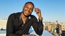 A man in a button-down shirt poses on a roof with a city skyline in the background.