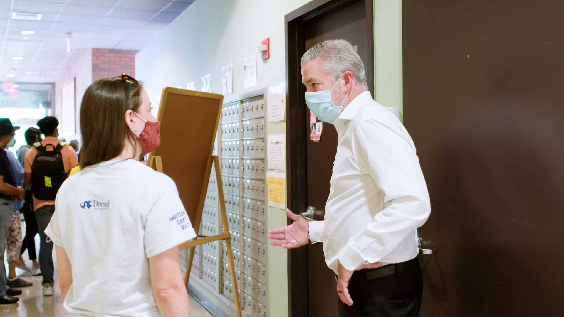 President John Fry answering a young woman’s inquiry during Student Move-In Day. They are standing in the hallway of a dormitory with other students and family nearby. Both are wearing masks.